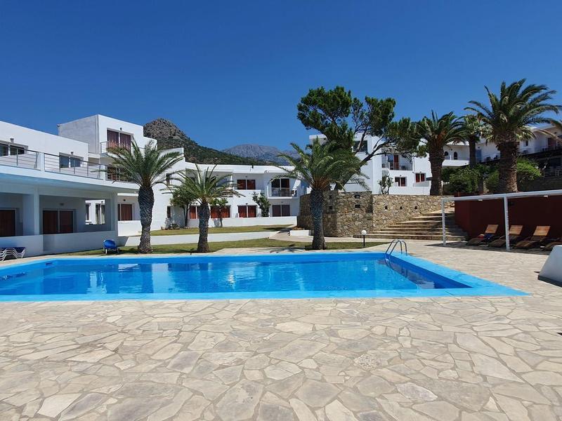 Área de piscina moderna en hotel con agua azul clara y edificios blancos bajo cielo azul.
