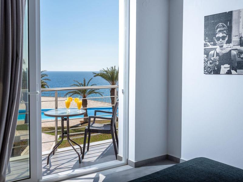 Vista desde una habitación de hotel con balcón hacia la piscina, el mar y palmeras en un día soleado.