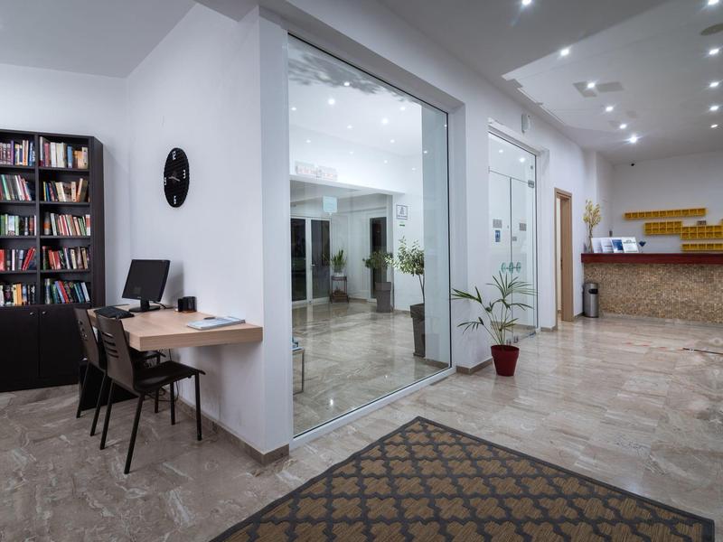 Recepción de hotel moderna con mostrador, escritorio con computadora y estantería cerca de la entrada.