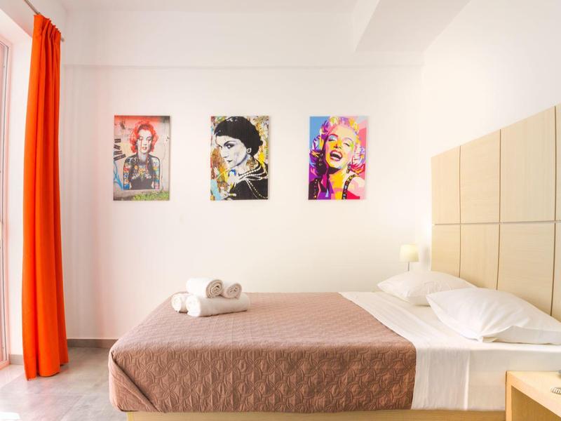 Habitación de hotel luminosa con cama grande, obras de arte en la pared y cortina naranja en la ventana del balcón.