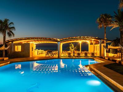 Piscina iluminada frente a un hotel por la noche con palmeras y agua azul clara.