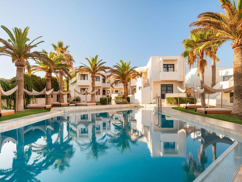Modern hotel pool with palm trees and white buildings under a clear blue sky.