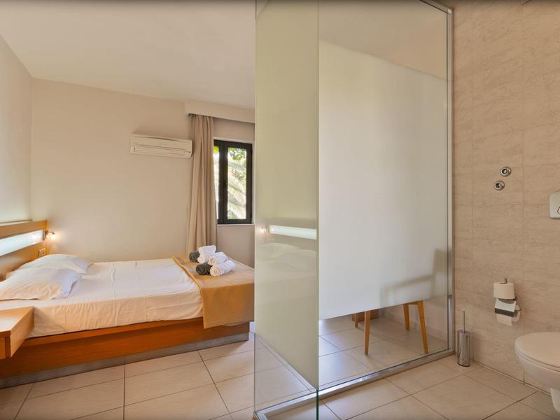 Bright hotel room with double bed and bathroom separated by glass wall.