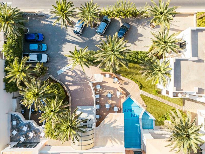 Aerial view of a hotel with pool, surrounded by palm trees and parked cars.