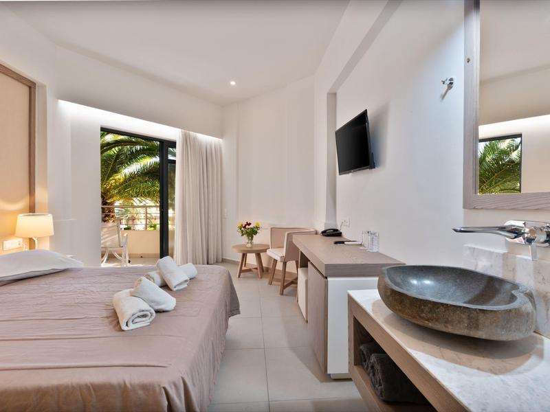 Brightly furnished hotel room with double bed, TV, washbasin, and balcony view with palm trees