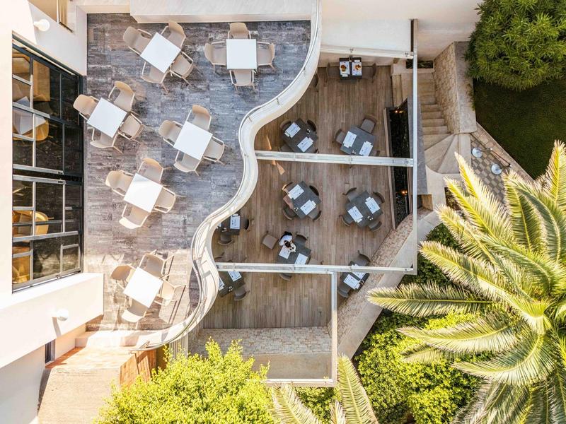 Bird's eye view of a terrace with seating areas, umbrellas, and palm trees in a hotel garden.