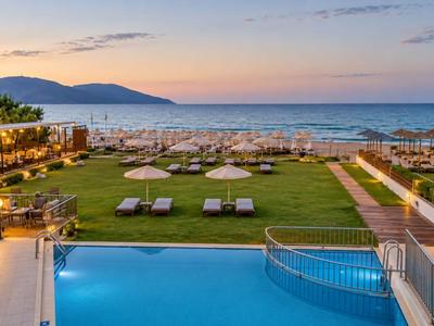 Piscine d'hôtel avec chaises longues, pelouse et plage sous un coucher de soleil coloré.