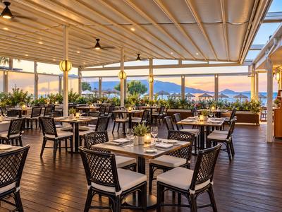 Restaurant d'hôtel ouvert avec sol en bois, tables dressées et vue sur les montagnes au coucher du soleil.