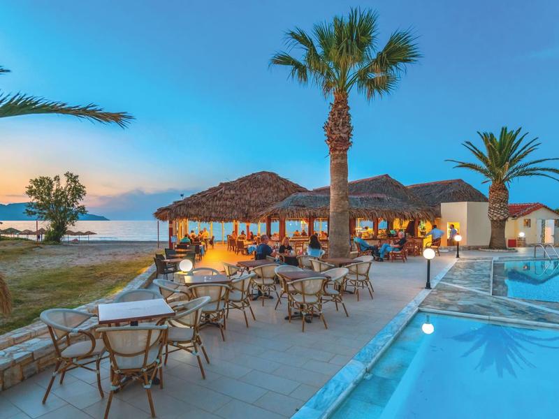 Terrasse d'hôtel en soirée avec piscine bordée de palmiers et vue sur la mer au coucher du soleil.