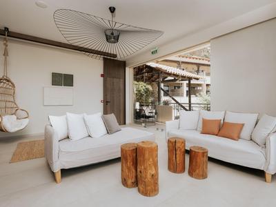 Área de estar luminosa con sofás blancos, taburetes de madera y una silla colgante junto a una puerta abierta.