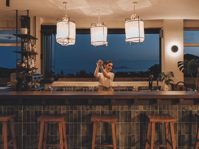 Barista che prepara cocktail in un bar d'hotel moderno con sgabelli e ampia finestra di notte.