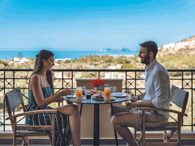Pareja disfrutando del desayuno en balcón con vista al mar bajo cielo despejado.