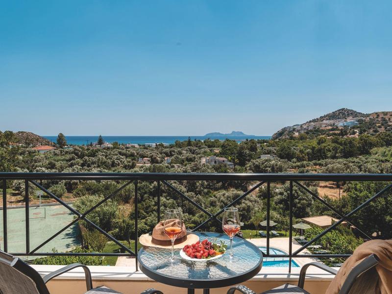 Balcone con tavolo e sedie, frutta e bevande, vista su paesaggio verde e mare.