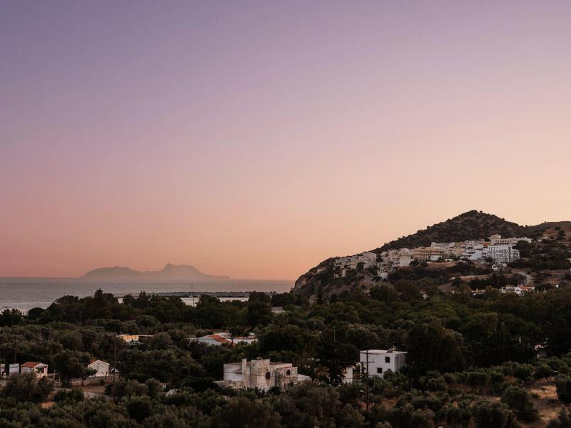 Vista di una città costiera al tramonto con montagne e mare sullo sfondo.