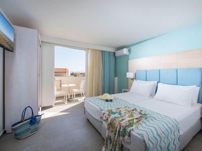 Habitación de hotel luminosa con cama doble, balcón y vista al mar, tonos azules suaves y decoración moderna.