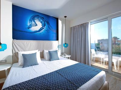 Habitación de hotel moderna con cama doble, acentos azules y balcón con vistas al exterior.