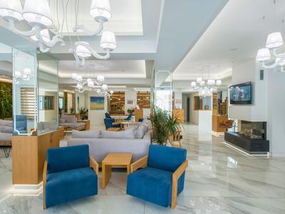 Área de recepción de hotel luminosa con sillones azules, sofás, detalles de madera y vidrio, iluminación moderna y suelo de mármol.
