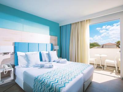 Habitación de hotel luminosa con cama doble, acentos azules y balcón con vista al cielo azul.