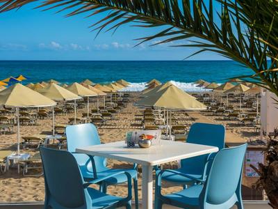 Blick auf einen Tisch mit Stühlen am Strand, viele Sonnenschirme und das Meer im Hintergrund.