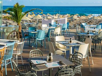 Offene Terrasse mit Tischen und Stühlen am Meer mit Sonnenschirmen im Hintergrund.