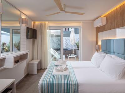 Helles Hotelzimmer mit Doppelbett, Deckenventilator und Blick auf eine Terrasse mit Pool.