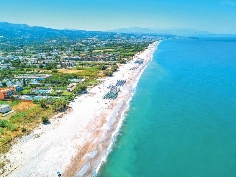 Larga playa de arena con agua turquesa, junto a paisaje verde y asentamientos bajo un cielo despejado.