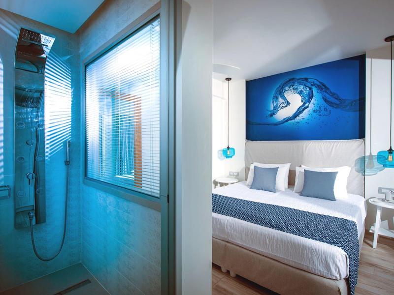 Habitación de hotel moderna con ropa de cama azul y ducha a ras de suelo con paredes de cristal.