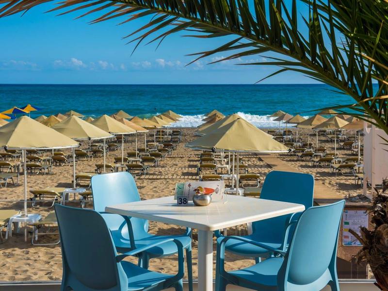 Playa con sombrillas y mar azul, frente a una mesa con sillas azules bajo hojas de palmera.