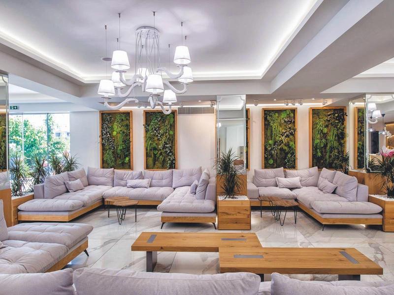 Sala de estar luminosa con grandes sofás, mesas de madera y plantas verdes en la pared.