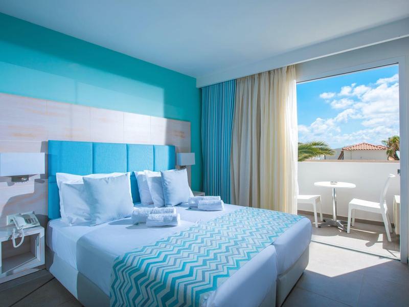Habitación de hotel luminosa con ropa de cama azul y blanca y balcón con vista al cielo.