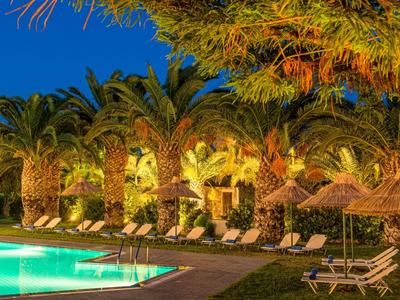 Illuminated pool area with lounge chairs, palm trees, and thatched umbrellas at dusk.