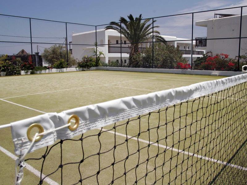 Un campo da tennis con una rete, circondato da edifici bianchi e palme sotto un cielo azzurro.