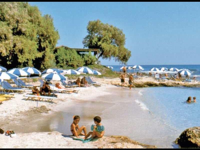 Spiaggia con ombrelloni, ospiti in acqua e riva rocciosa sotto il cielo azzurro.