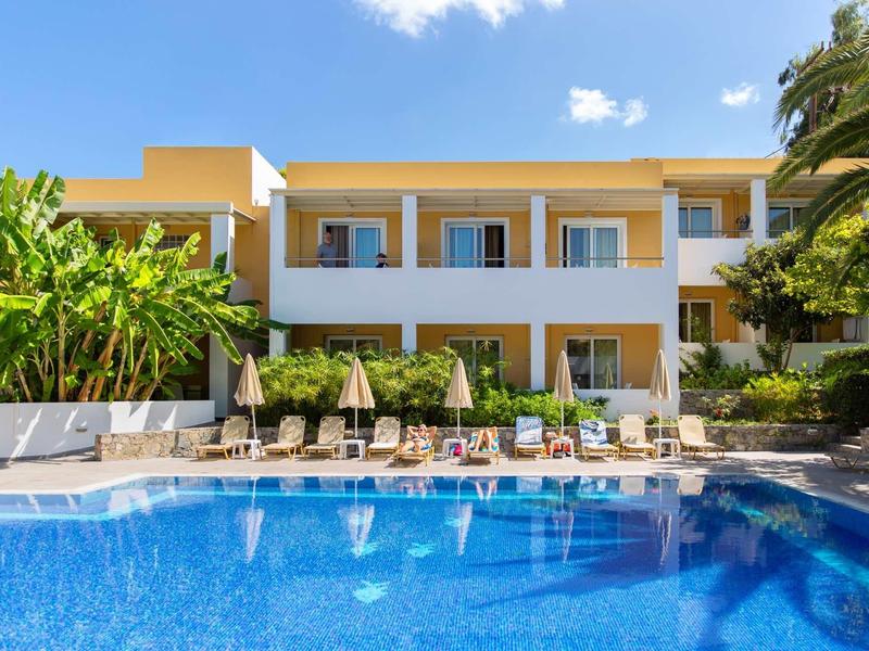 Hôtel avec terrasse ensoleillée et piscine bleue sous un ciel clair, entouré de palmiers.