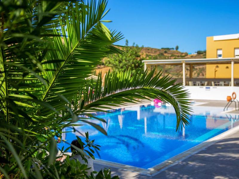 Piscine bleue à côté d'un bâtiment d'hôtel jaune avec des palmiers au premier plan sous un ciel bleu.