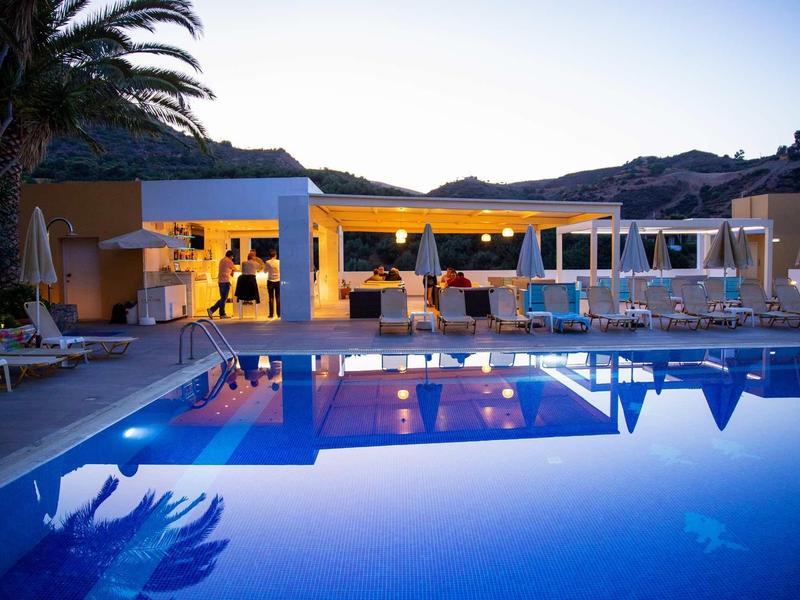 Piscine en soirée avec hôtel éclairé, coins salons et palmiers.
