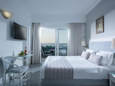 Modern hotel room with double bed, balcony, and sea view in bright daylight.