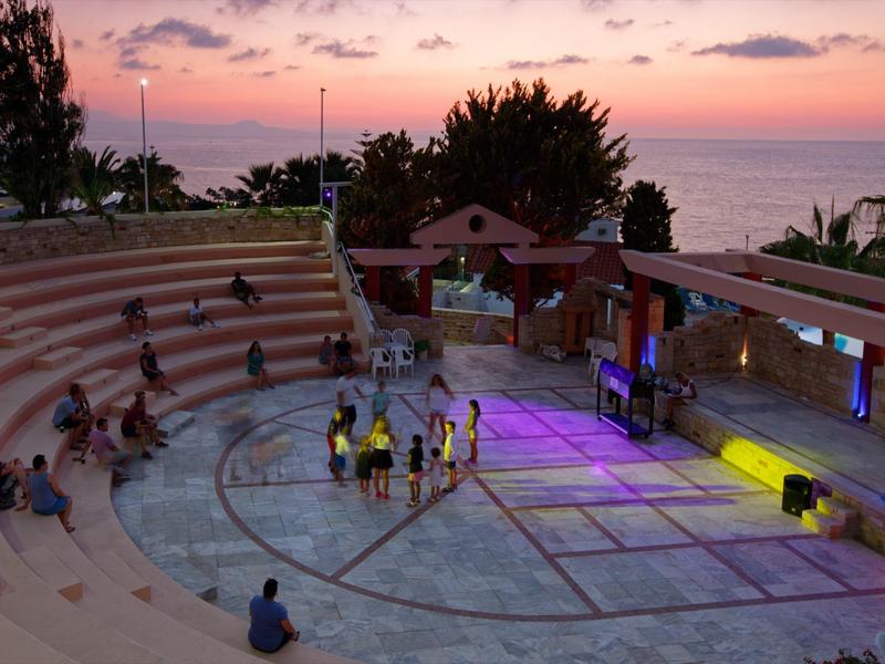 Outdoor amphitheater at sunset by the sea with audience and trees in the background.
