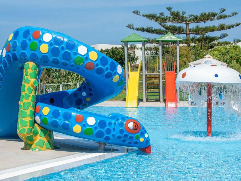 Colorful water slide and water sprinkler at an outdoor pool with surrounding plants.