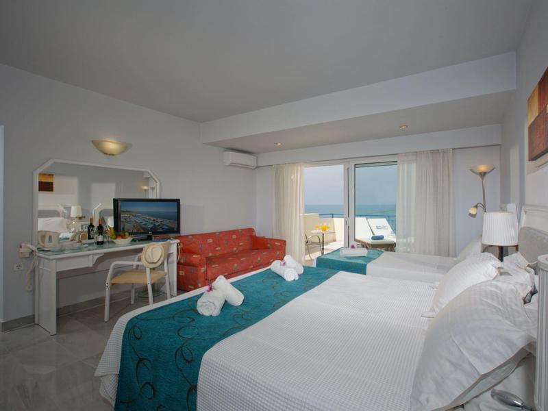 Bright hotel room with double bed, TV, desk, and balcony with sea view.