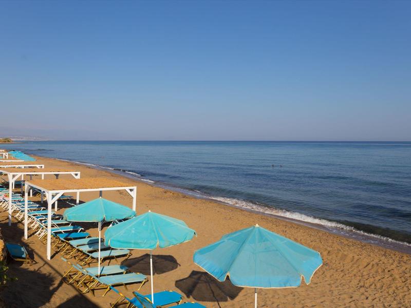 Sandy beach with blue umbrellas and wooden canopies by the calm sea under a clear blue sky.