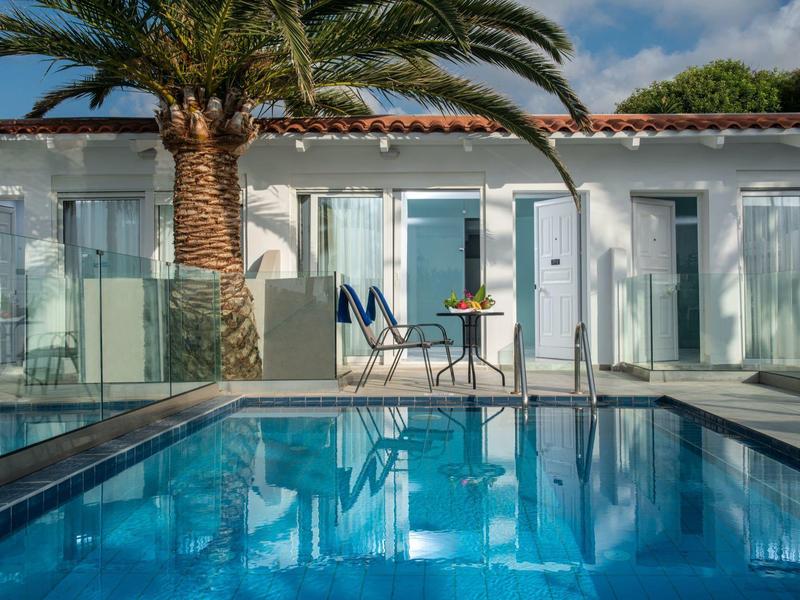 Modern pool with palm trees in front of white hotel rooms and patio furniture in clear weather.