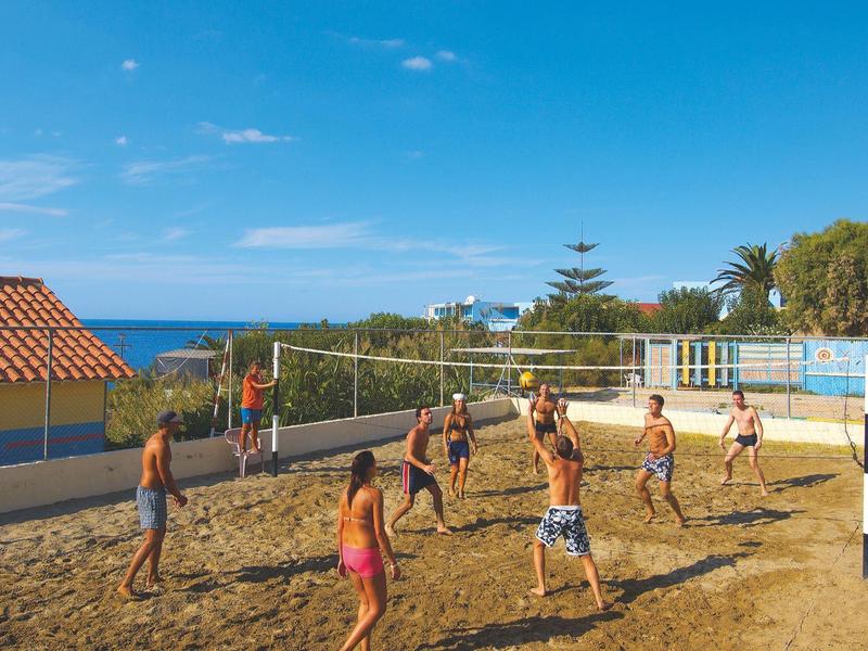 Group of people playing volleyball on sand beside a hotel with sea view.
