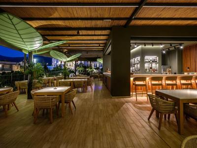 Ristorante aperto con pavimento in legno, tavoli e sedie sotto grandi paralumi verdi alla luce della sera.