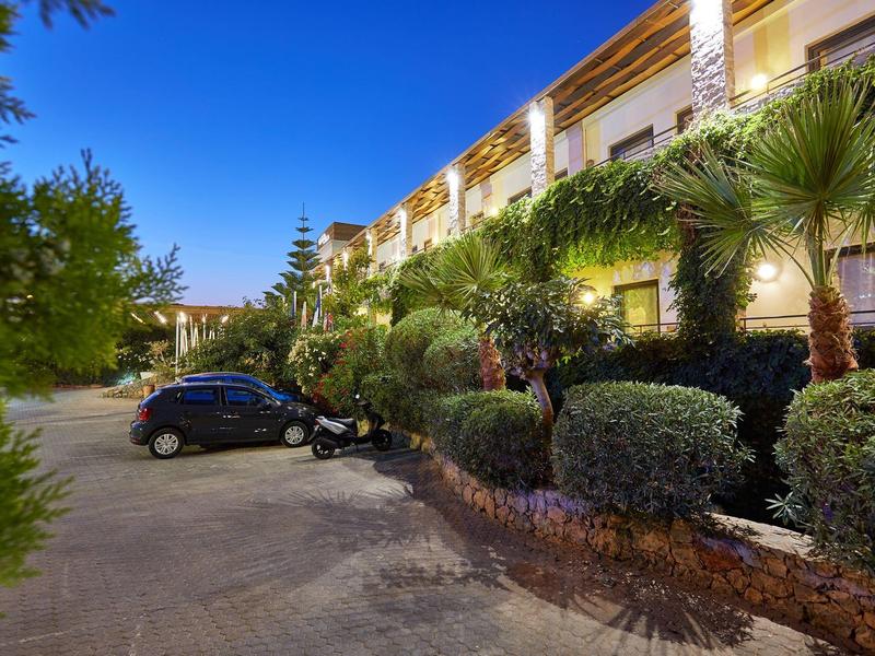 Esterno di un hotel con vialetto, auto parcheggiate e camere illuminate al crepuscolo.