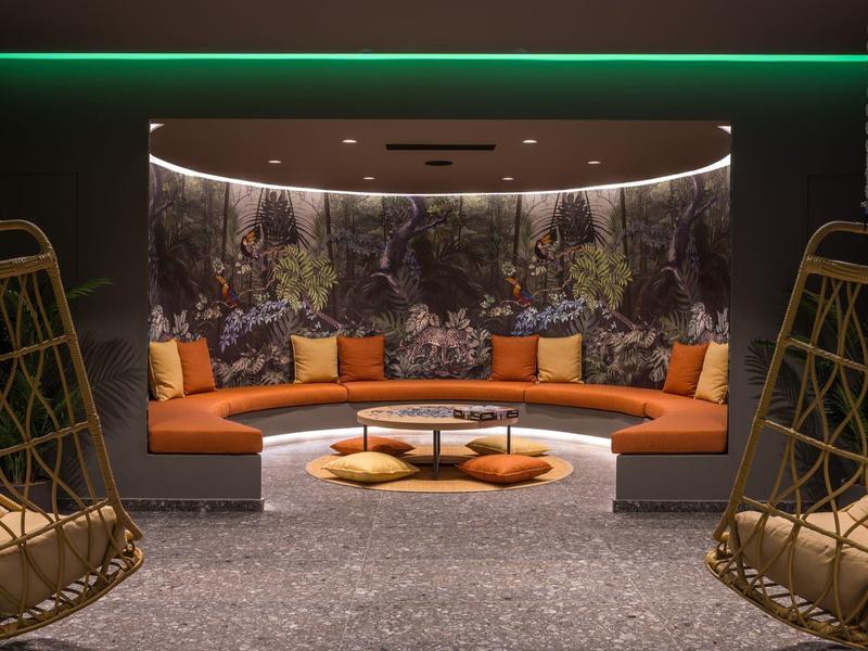 Zona salotto rotonda con cuscini arancioni, sedie sospese e carta da parati della foresta in ambiente accogliente.