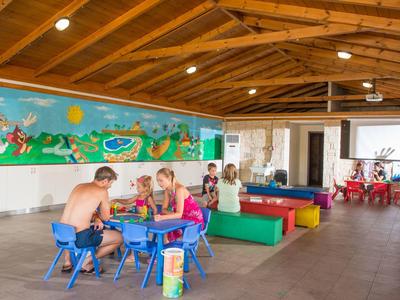 Bambini giocano e fanno lavoretti a piccoli tavoli in una sala colorata e aperta con soffitto in legno.