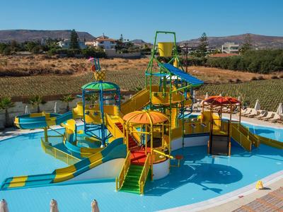 Area giochi acquatica colorata con scivoli in una piscina, circondata da lettini e montagne sullo sfondo.