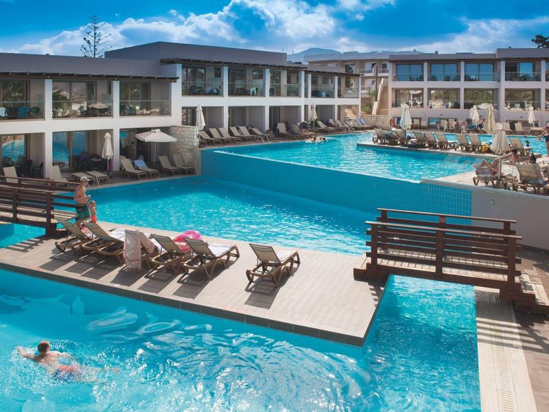 Resort moderno con grande piscina all'aperto, lettini e edifici su due piani sotto un cielo azzurro.