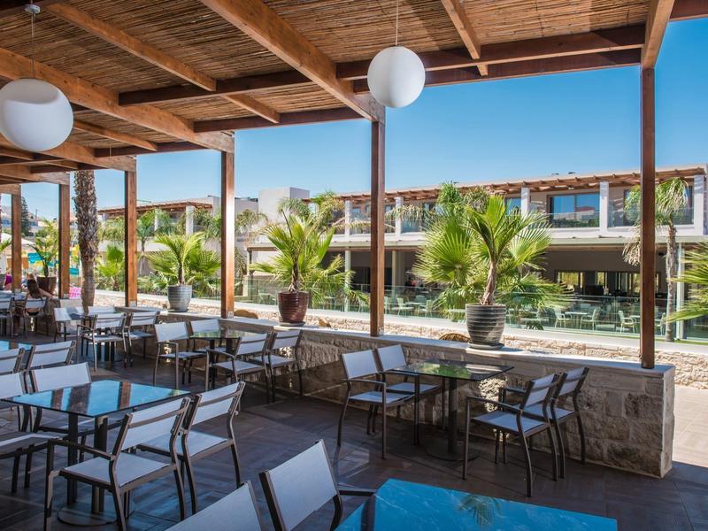 Terrazza esterna coperta con tavoli e sedie, vista su edifici dell'hotel e palme.